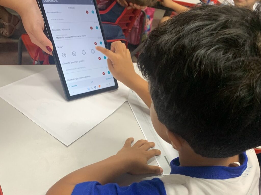 Criança participando de um teste de aceitabilidade de escala hedônica presente em um tablet. A imagem retrata a importância de realizar testes de aceitabilidade nas escolas.