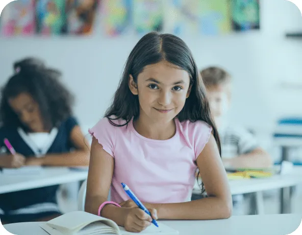 Menina sorridente sentada em uma sala de aula, segurando um lápis e olhando para a câmera, com colegas estudando ao fundo, destacando o ambiente educacional e a aprendizagem escolar.